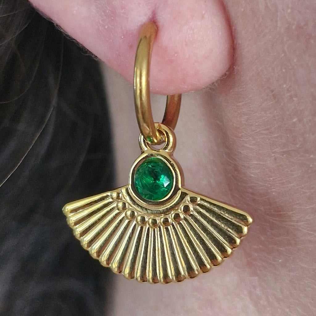 A gold hoop earring with a dangling fan-shaped charm and a round green gemstone, shown worn on a person's ear.