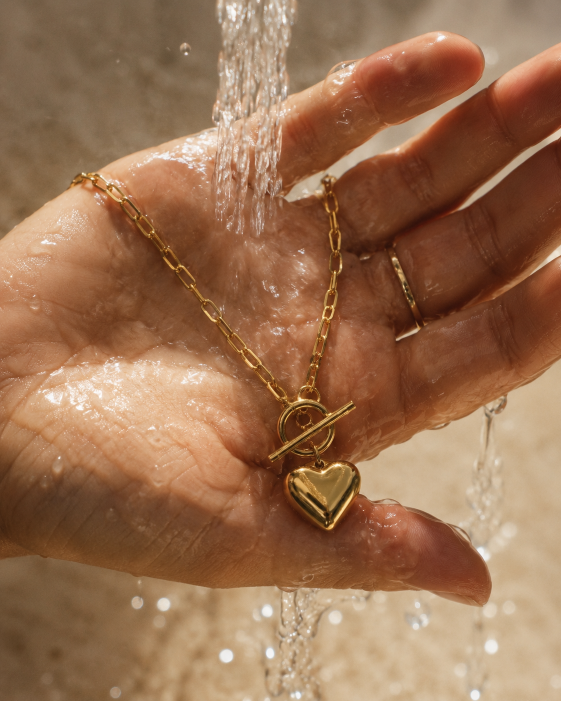 A gold chain necklace with a toggle clasp and heart pendant is held in a person's hand under running water. The necklace has a polished gold finish, and the hand is wet with water droplets, with a neutral beige background.