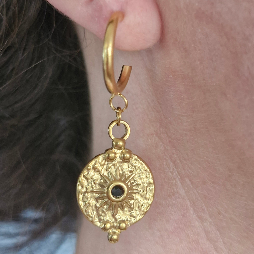 A close-up of a gold huggie hoop earring with a dangling textured circular charm featuring a central black eye detail, shown worn on a person's ear.