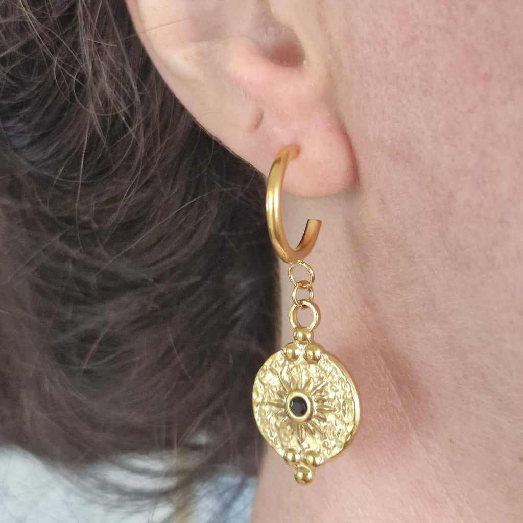 A close-up of a person's ear wearing a gold huggie hoop earring with a dangling textured circular charm featuring a small black center detail.