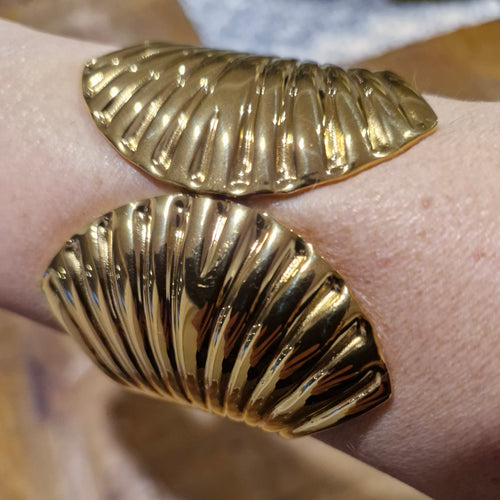 A close-up of a person's wrist wearing a wide, sculptural gold cuff bracelet with two large, ribbed shell shapes.