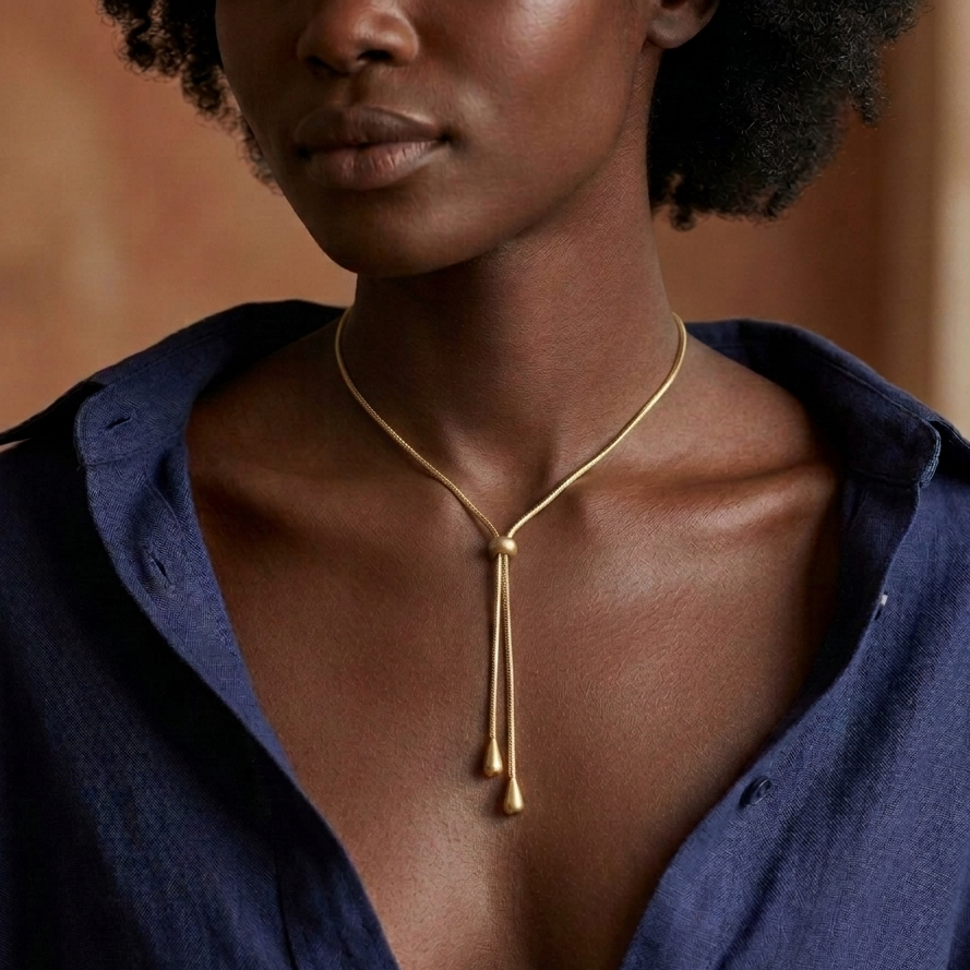 A close-up of a model wearing a delicate gold chain lariat necklace with two dangling teardrop charms, styled with an open navy blue shirt.