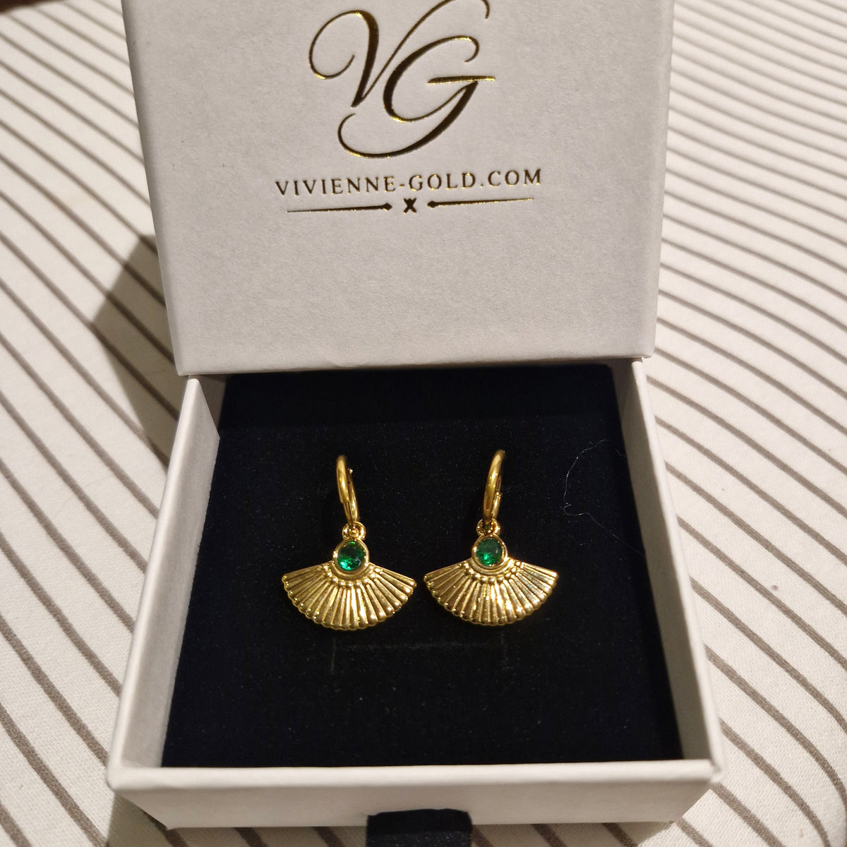 A pair of gold fan-shaped drop earrings with small green stone centers, presented in an open white jewelry box with the brand name VIVIENNE-GOLD.COM printed inside the lid, set on a striped fabric surface.