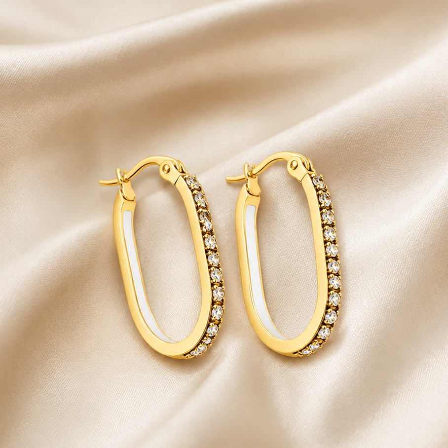 A pair of gold hoop earrings with a row of clear crystals along one side, displayed on a draped beige satin fabric background.