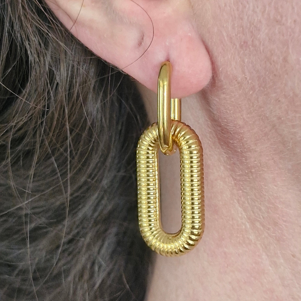 A close-up of a person's ear wearing a textured gold hoop earring with a large, vertically oriented open oval drop.