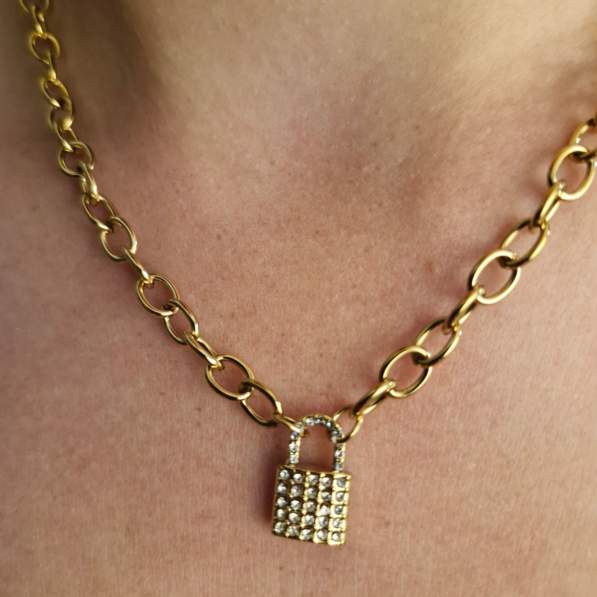 A gold chain necklace with a small padlock charm encrusted with clear rhinestones, shown worn on a person's ankle.