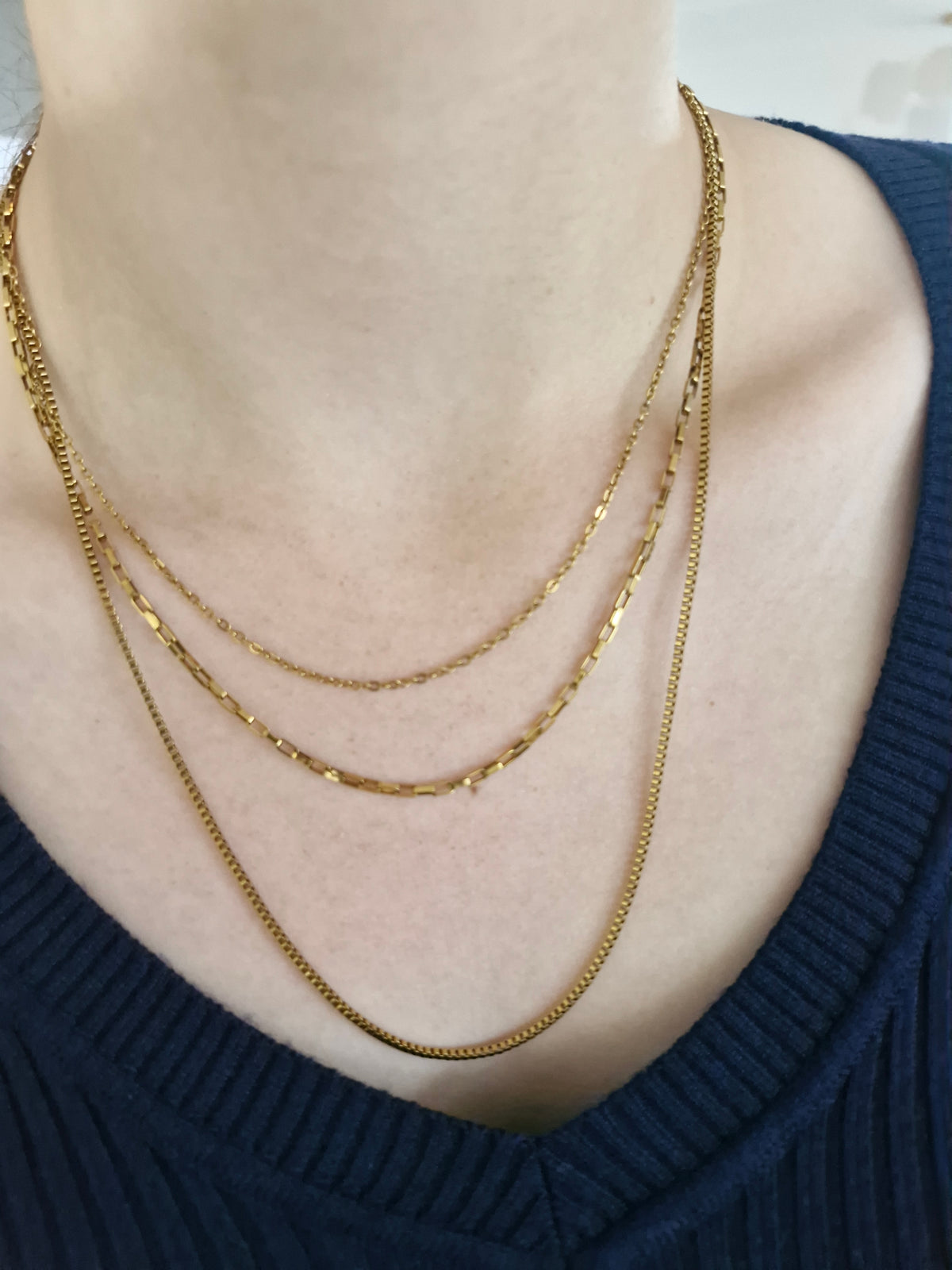 A close-up of a person's neck and upper chest wearing three layered Maggie gold-tone chain necklaces over a dark navy ribbed V-neck top.
