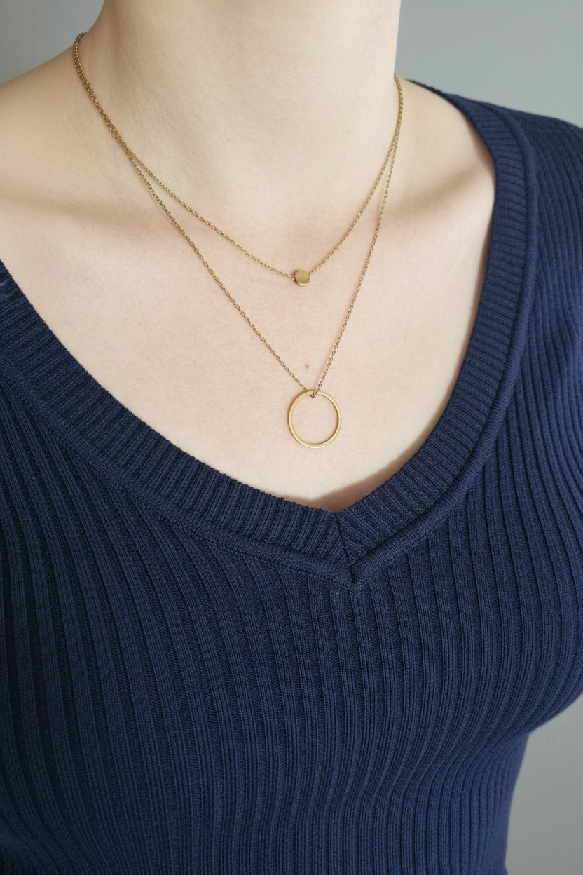 A close-up of a person wearing a double-layered gold necklace. The necklace features a delicate chain with a small flat circular disc pendant on the top layer and a longer chain with a larger open circle pendant on the bottom layer.
