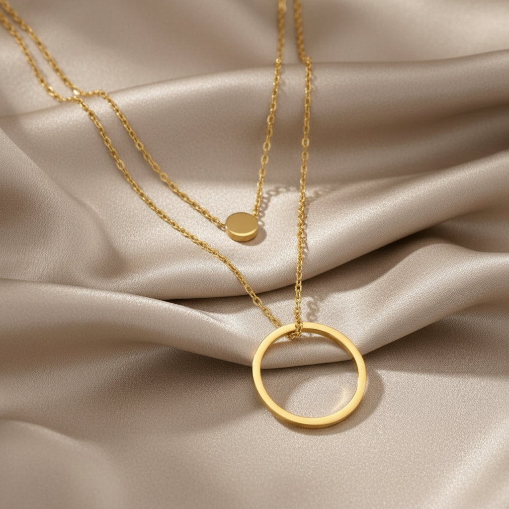 A gold layered necklace set displayed on a textured beige surface. The set features a longer chain with a large open circular pendant and a shorter chain with a small flat circular disc pendant.