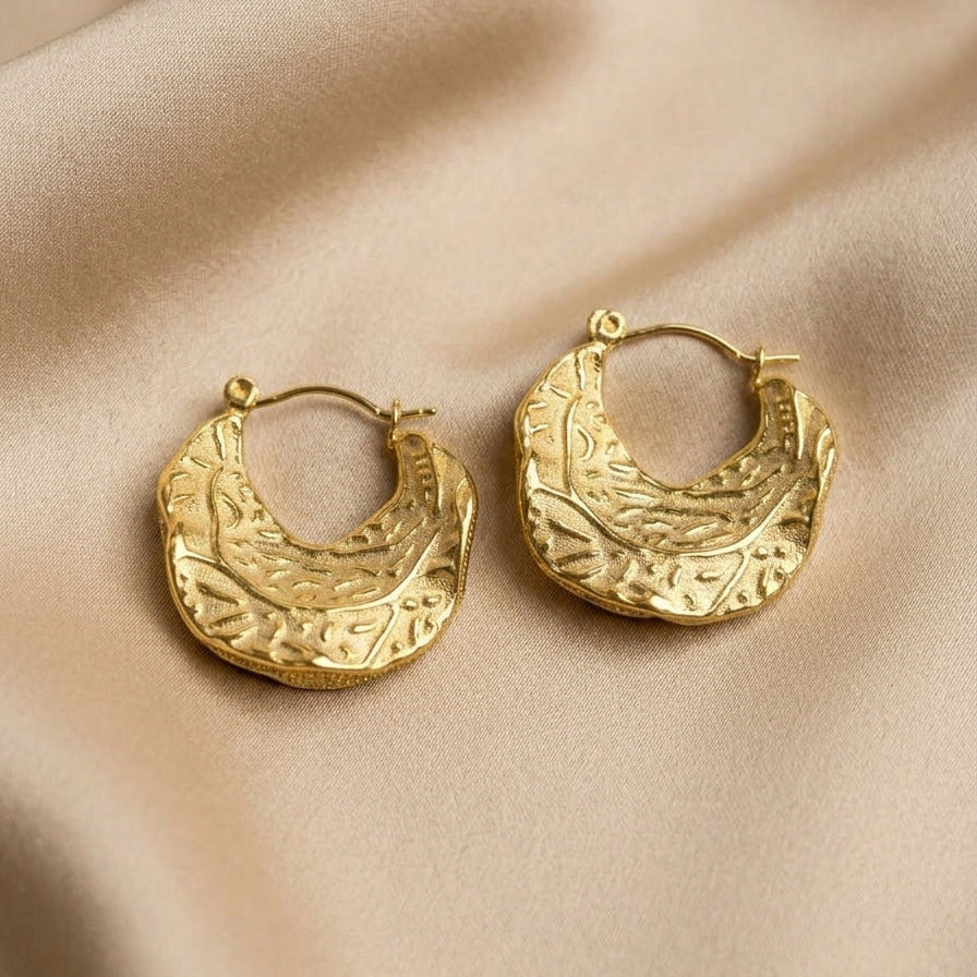 A pair of textured gold crescent-shaped hoop earrings resting on draped beige satin fabric.