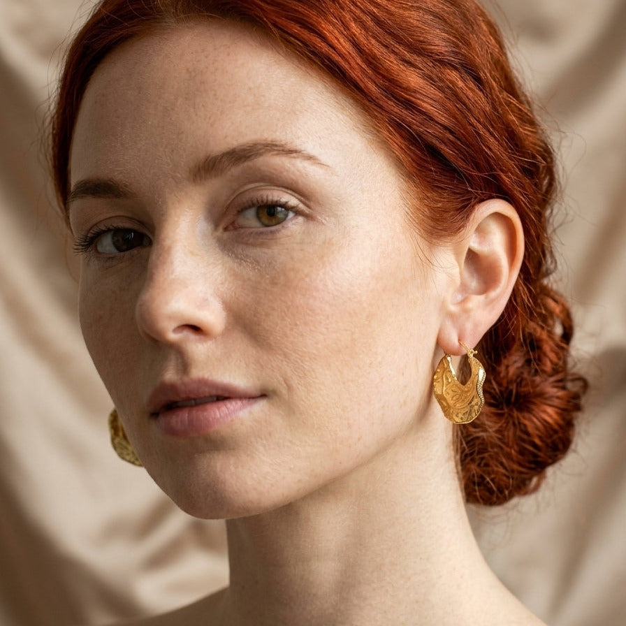 A close-up portrait of a woman with red hair wearing textured gold hoop earrings against a soft beige fabric background.