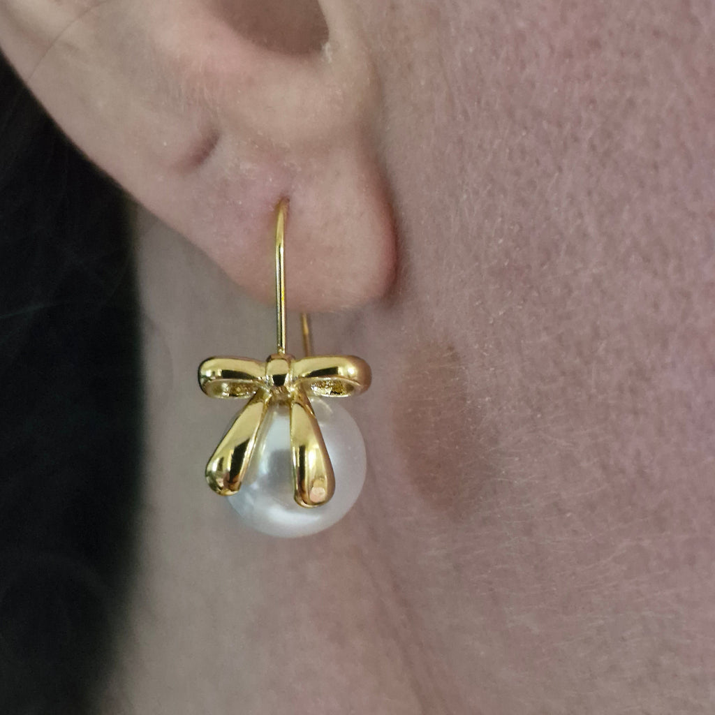A close-up of a person's ear wearing a gold bow-shaped hoop earring with a white pearl drop.