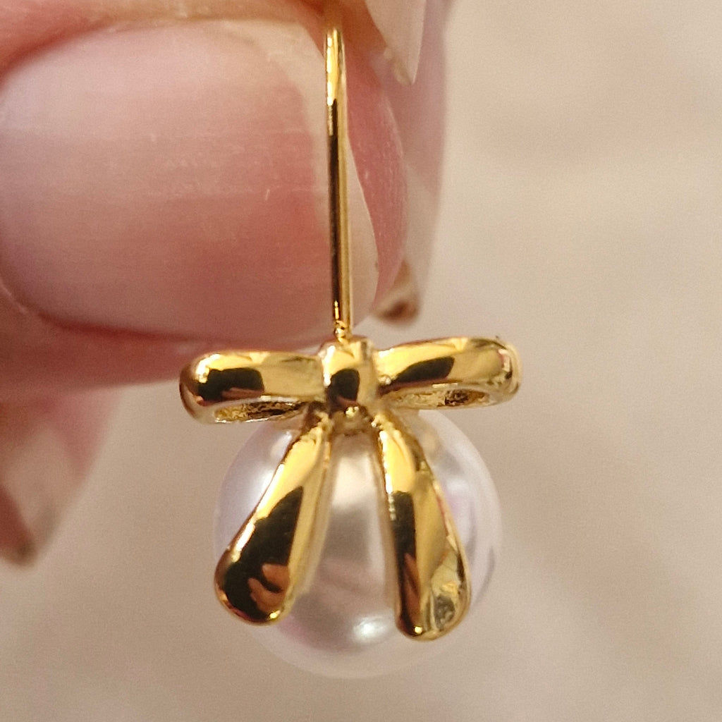 A close-up of a hand holding a gold bow-shaped earring with a white pearl pendant.
