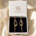 A pair of gold teardrop-shaped drop earrings displayed inside an open white jewelry box with a black velvet insert. The inside lid of the box shows the brand name 'Vivienne Gold' in gold lettering.