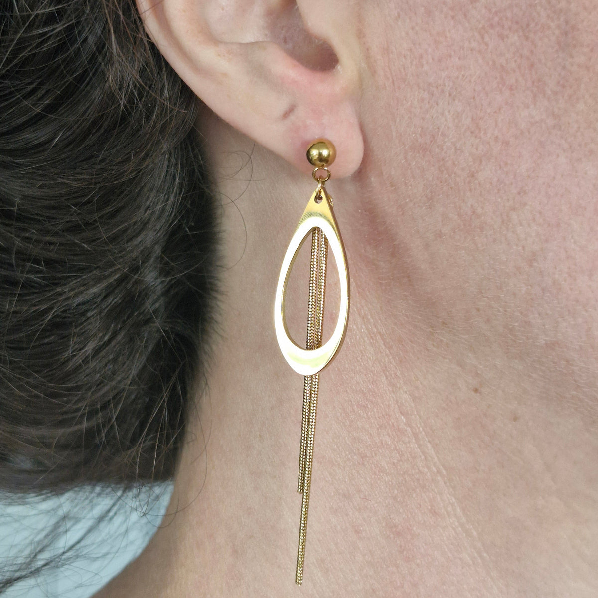 A close-up of a person's ear wearing a gold dangle earring with an oval cutout design and thin chain-like drops.