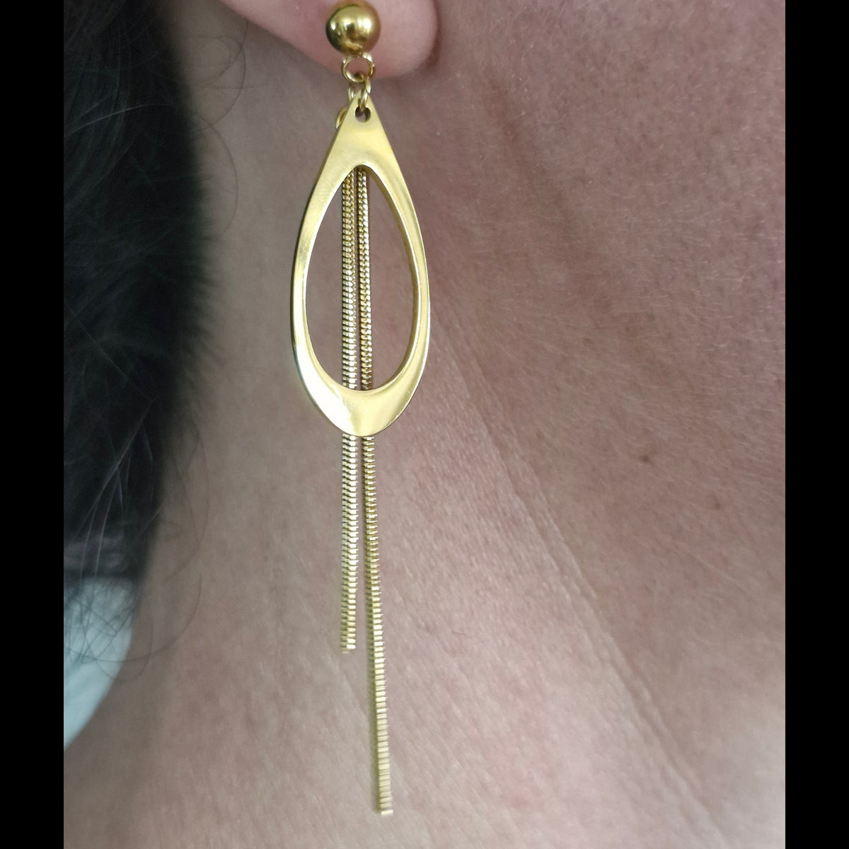 A close-up of a gold dangle earring worn on a person's ear, featuring a polished oval frame with two hanging textured chain strands.