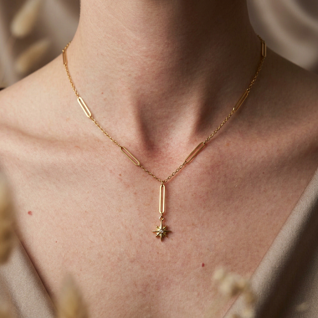 A delicate gold chain necklace with multiple bar links and a small starburst charm, shown worn on a person's neck over a white tank top.