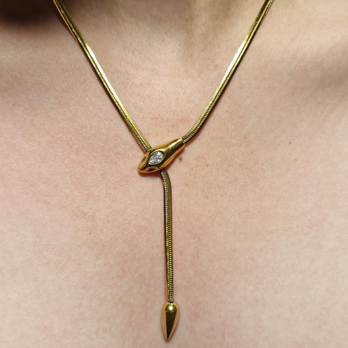 A gold snake chain anklet with a central teardrop-shaped connector set with a small clear gemstone, featuring a pointed tip on one end, shown worn on skin.