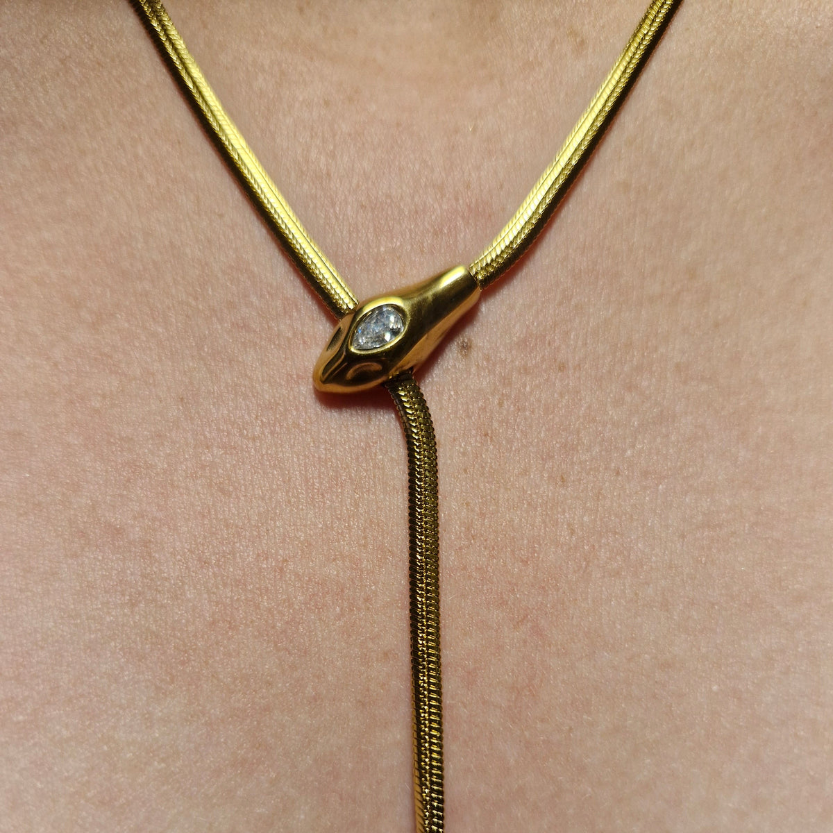 A close-up of a gold necklace with a Y-shaped design, featuring a small clear gemstone in the center and a textured snake chain leading to pointed ends, shown on a beige skin background.