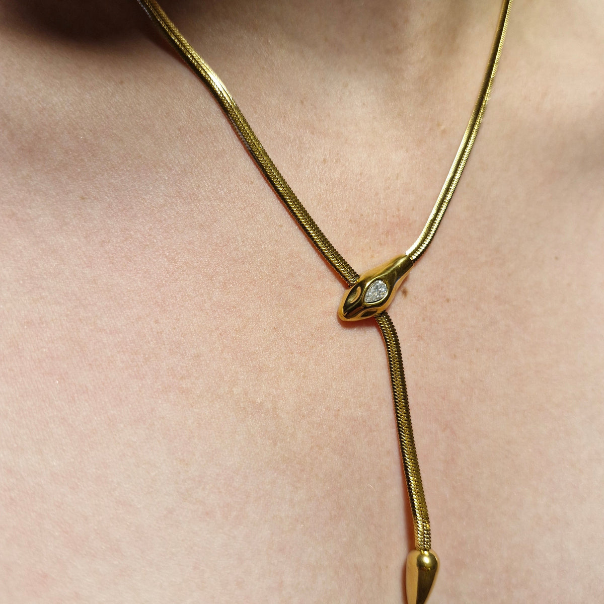 A close-up of a gold snake pendant necklace worn on a person's neck, featuring a diamond-set head and a spear-shaped dangling tip.