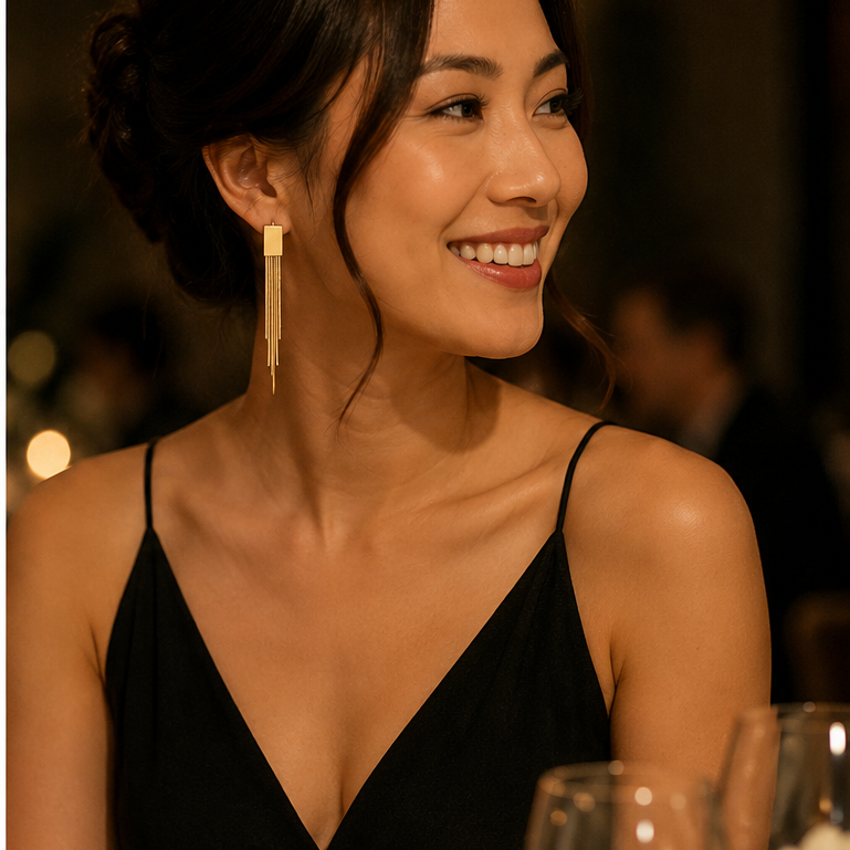 A woman with dark hair in an updo wearing a black spaghetti-strap dress and a long, rectangular gold dangle earring, smiling in a warmly lit social setting.