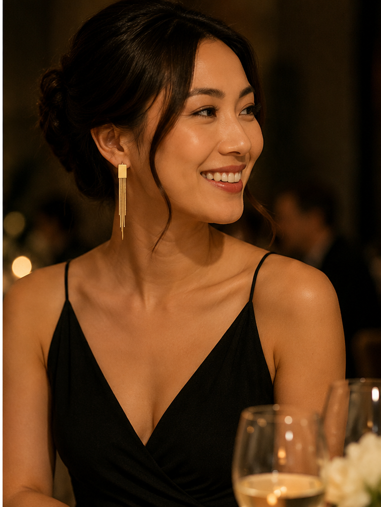 A woman with dark hair in an updo wearing a black spaghetti-strap dress and a long, rectangular gold dangle earring, smiling in a warmly lit social setting.